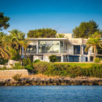 A modern luxurious seafront property located in Mallorca, Spain, with contemporary design, big glass windows. green garden with palm trees and the sea in front on a sunny day